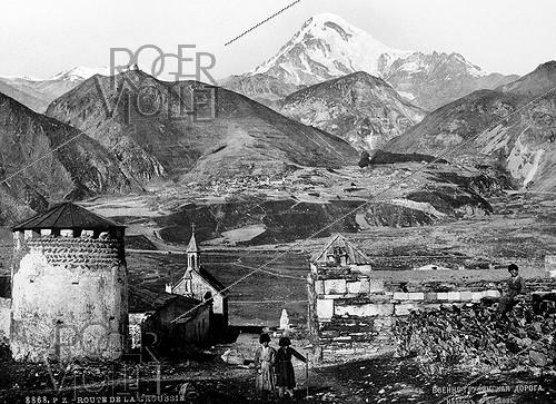 Kazbegui (Géorgie). Le village (au premier plan) et l'ancienne route militaire avec le Kazbek (5033 mètres) et, à gauche, l'église de Gergeti Tsminda Sameba (église de la Sainte-Trinité, XIVème siècle) et son clocher. Fin XIXème-début XXème siècles.